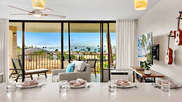 Ein geräumiges Zimmer mit einer großen Schiebeglasdoor, die auf einen Balkon mit Blick aufs Meer und Palmen führt. Ein Esstisch ist gedeckt, mit Tellern und Gläsern, und an der Wand hängt eine Gitarre.