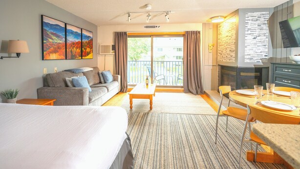 A hotel room with a bed, a sofa, a dining table with chairs, a TV, and a balcony with a view of the outside. - Ski-in, Ski-out Studio Condo at Smugglers' Notch Resort (Cambridge)
