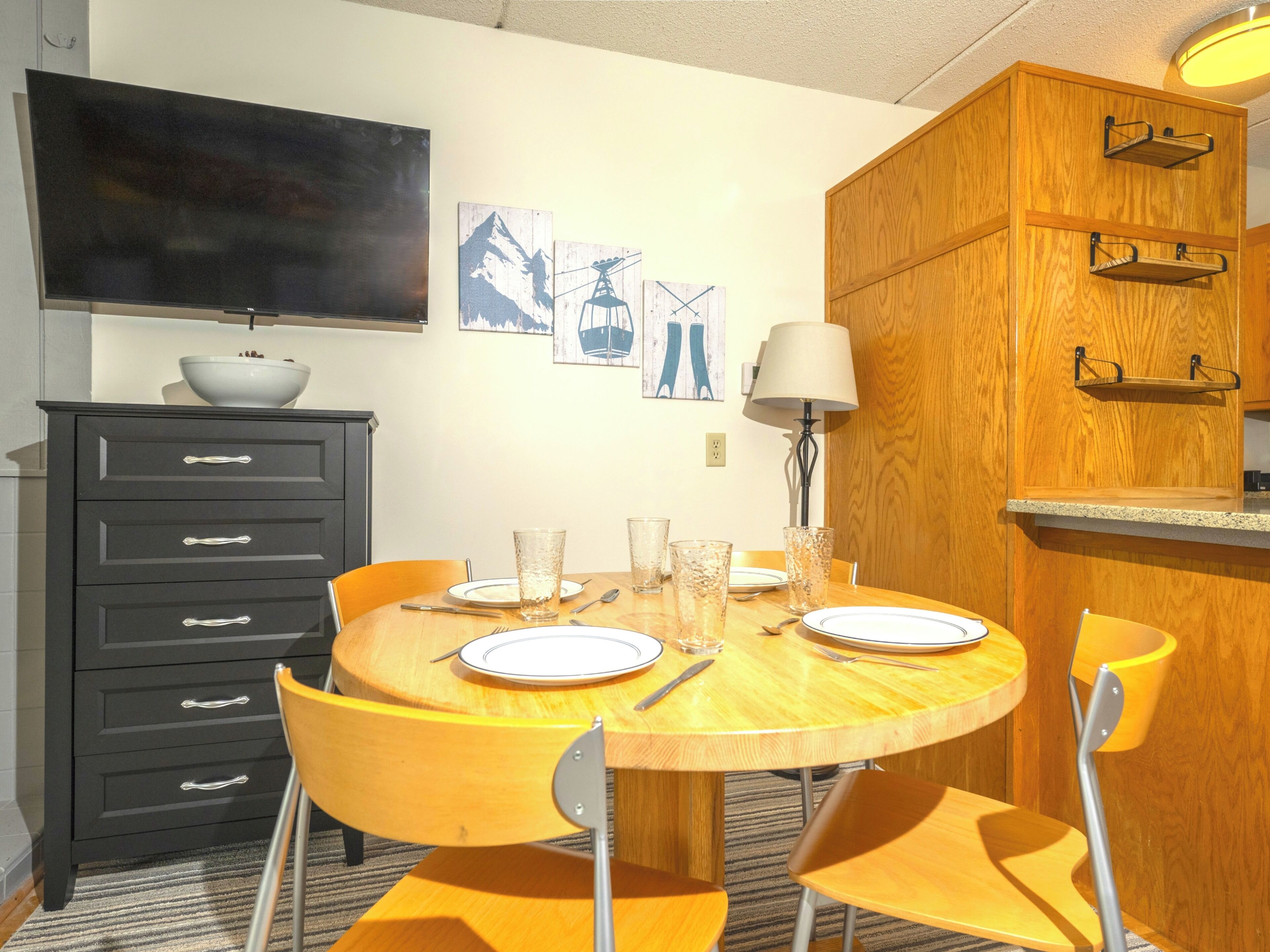 Un coin repas avec une table ronde en bois, quatre chaises, un téléviseur placé au-dessus de la table et des œuvres d’art accrochées au mur.