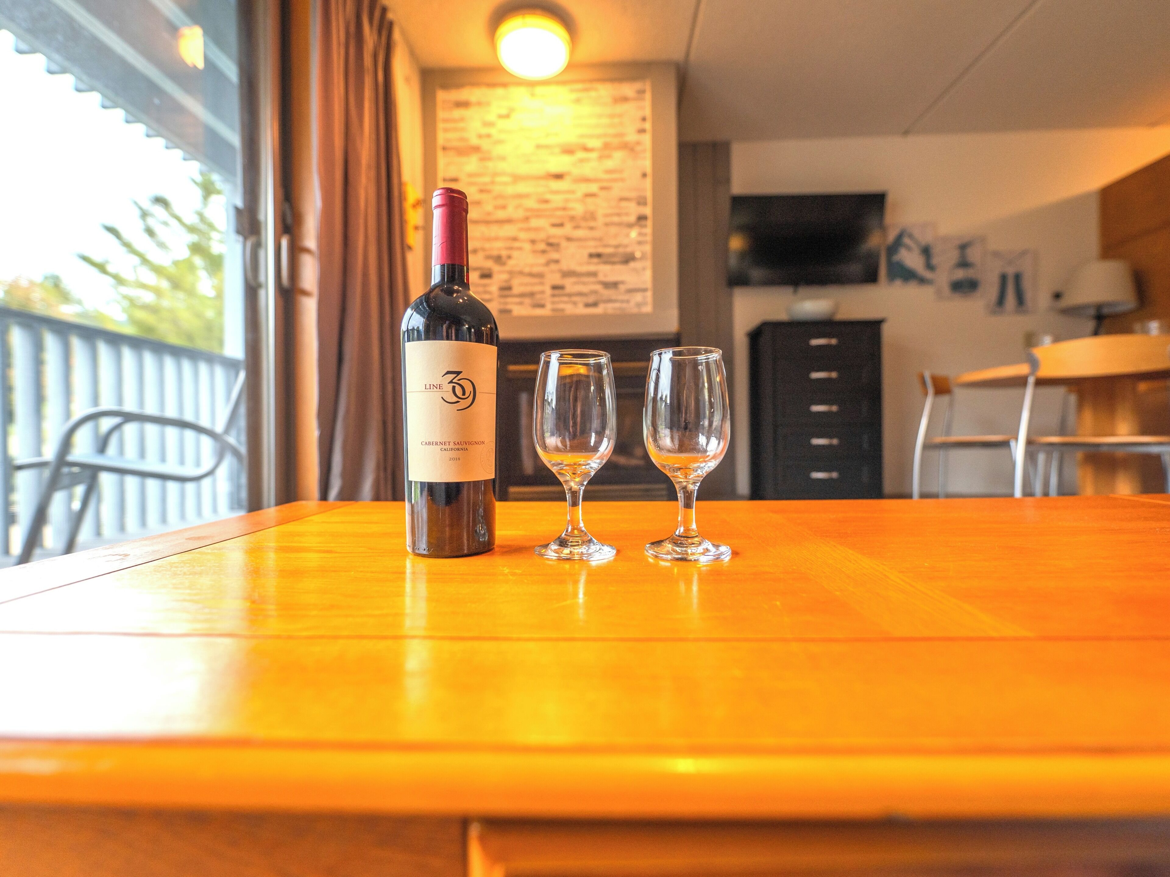 Une bouteille de vin et deux verres sur une table en bois, dans une pièce où se trouvent une télévision et un mur décoré d’œuvres d’art.