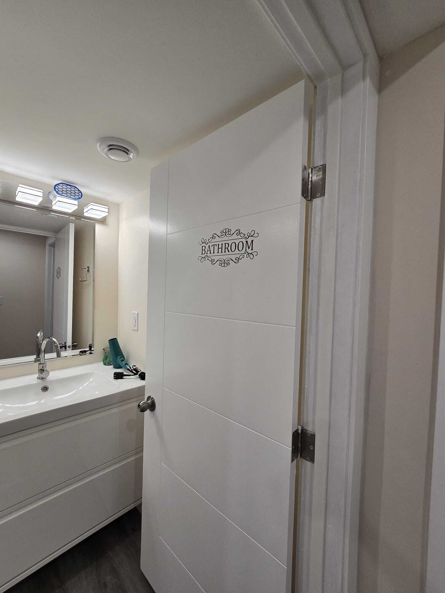 Un baño con puerta blanca, espejo, lavamanos y toallero.