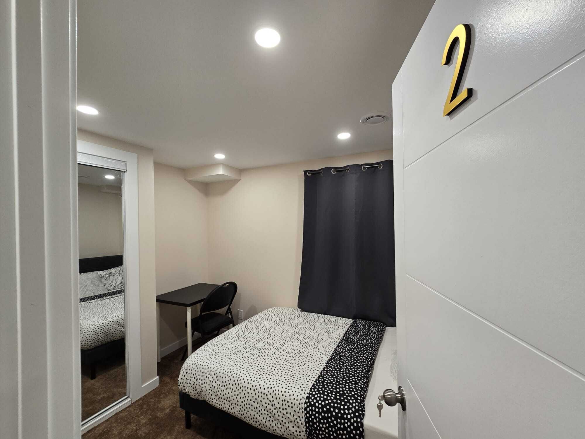 Ein kleiner, gut beleuchteter Raum mit einem Bett, einem Schreibtisch und einem Stuhl. Ein schwarzer Vorhang ist teilweise heruntergezogen, und auf der Wand ist die Nummer 2 zu sehen.