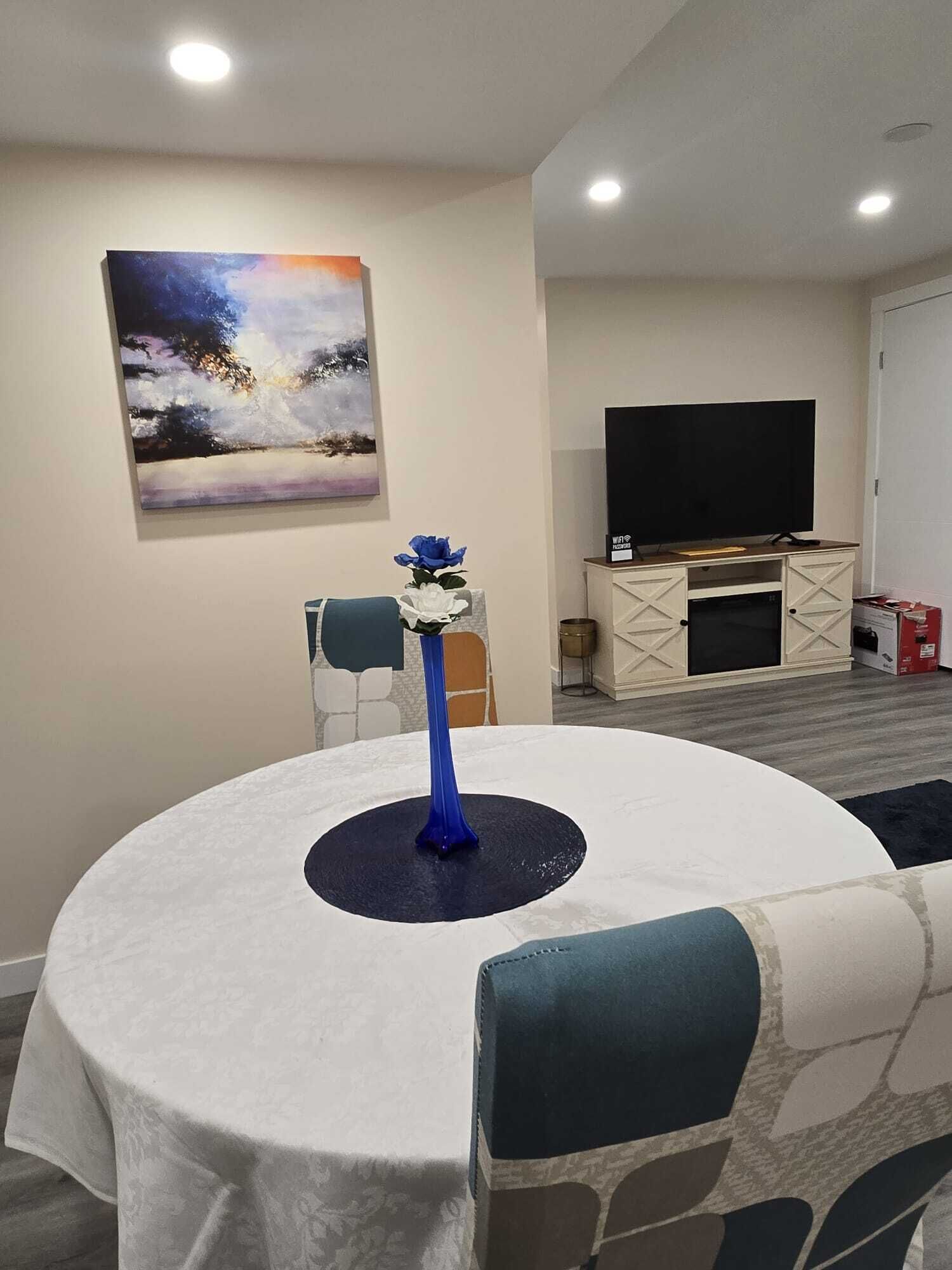 Un comedor con una mesa redonda, sillas, un florero con flores, un cuadro en la pared y un televisor sobre un mueble.