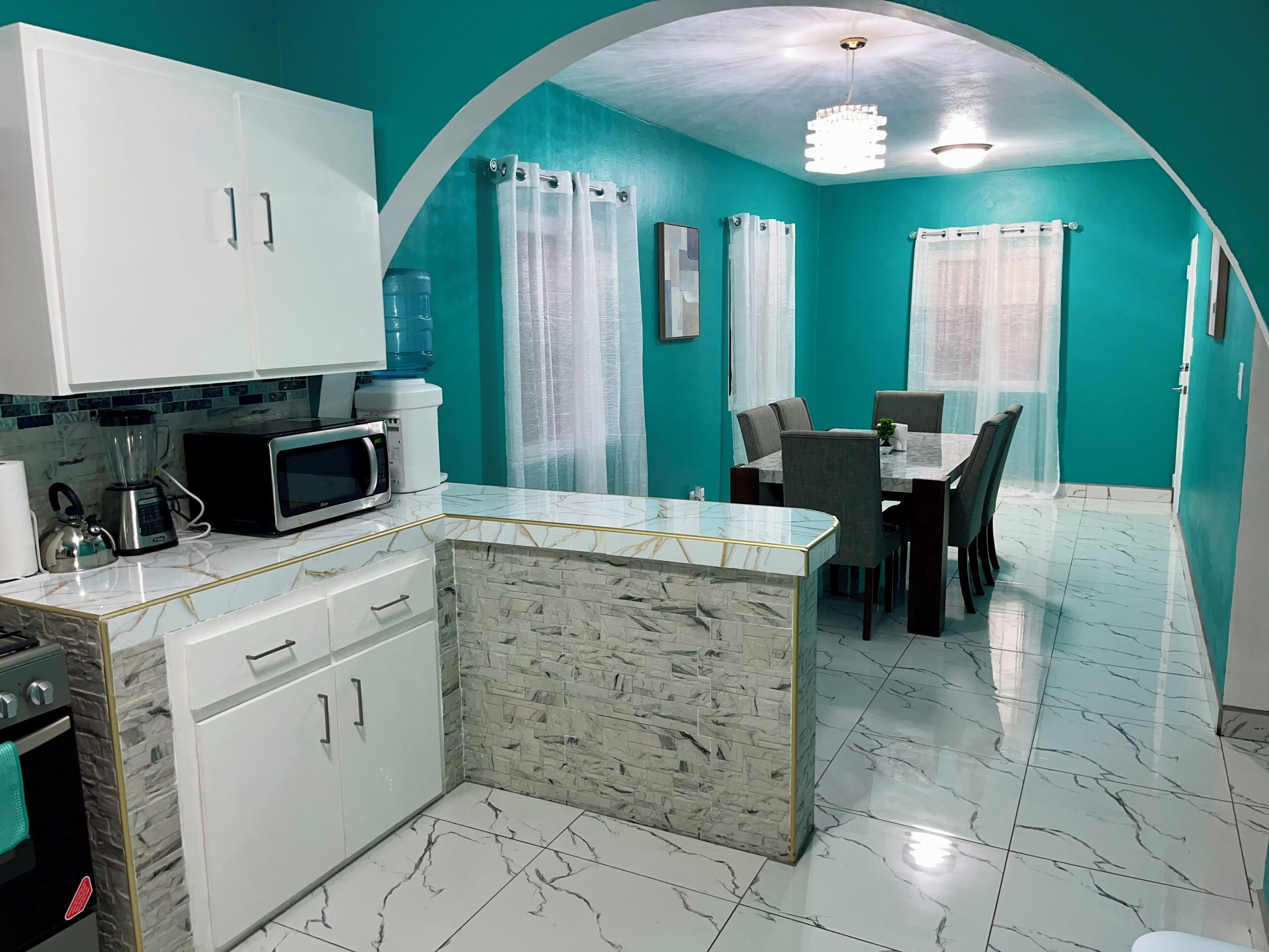 A kitchen with white cabinets, a microwave, a water dispenser, and a dining area with a table and chairs.