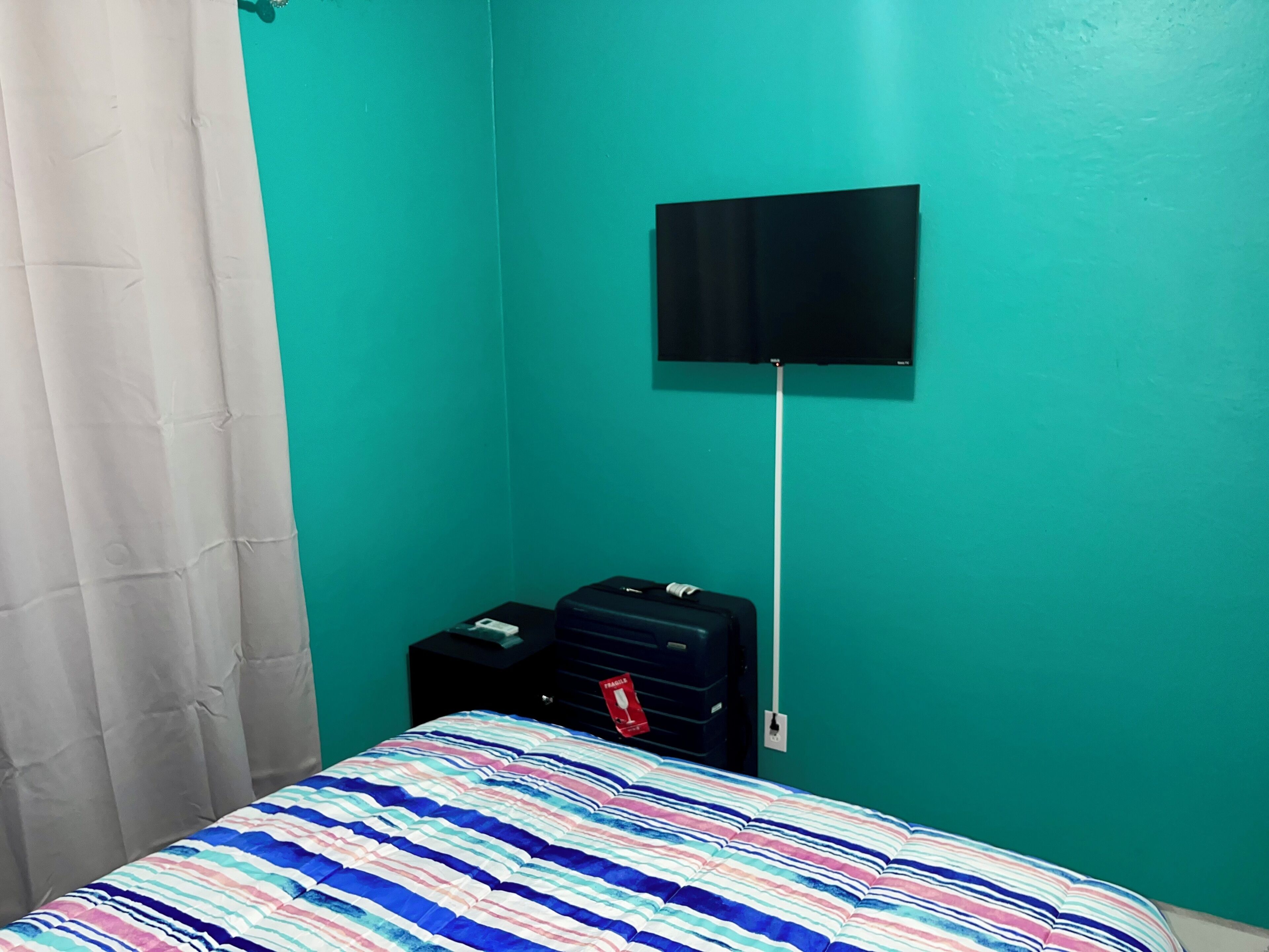 Una habitación con paredes color esmeralda, una cama con cubrecama a rayas, una maleta negra sobre una mesita y un televisor montado en la pared.