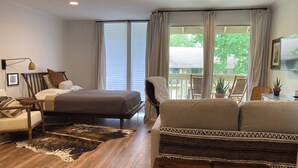Ein Schlafzimmer mit einem Bett, einem Sessel, einer Couch, Holzboden und einem großen Fenster mit Vorhängen.