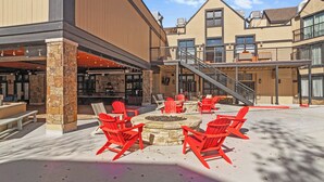 An outdoor seating area with red Adirondack chairs and a fire pit, surrounded by a building with a stone pillar and a staircase leading to an upper level. - Carriage House 101 | Kitchenette | Near Dining & Shopping | Hot Tub | Pool (Park City)