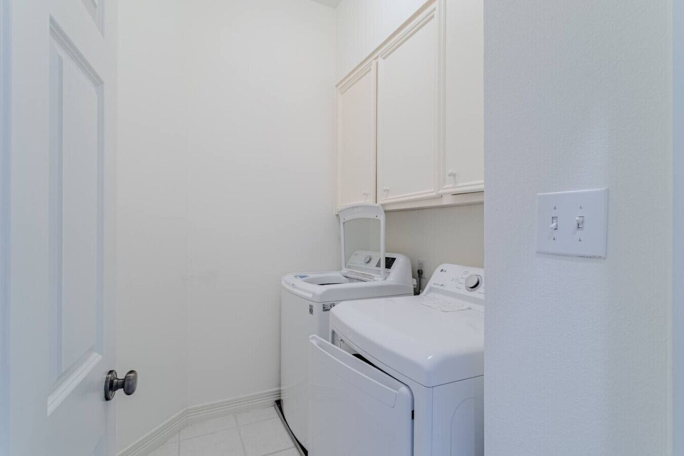 Uma lavanderia com duas máquinas de lavar roupa brancas e uma secadora, paredes brancas e uma porta.