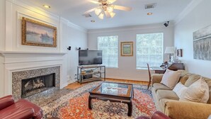 A living room with a fireplace, a television on a stand, a ceiling fan, and a large window with blinds. - Cozy Townhouse near Midtown (Houston)