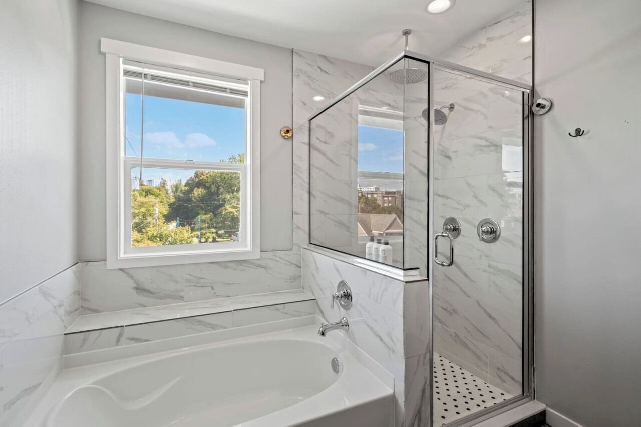 A modern bathroom with a glass-enclosed shower, a large bathtub, and a window offering a view of greenery and buildings.