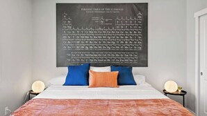 Un dormitori amb un gran tauler periòdic dels elements a la paret, un llit amb almoinades blaves i taronja, i taules de nit amb làmpades.