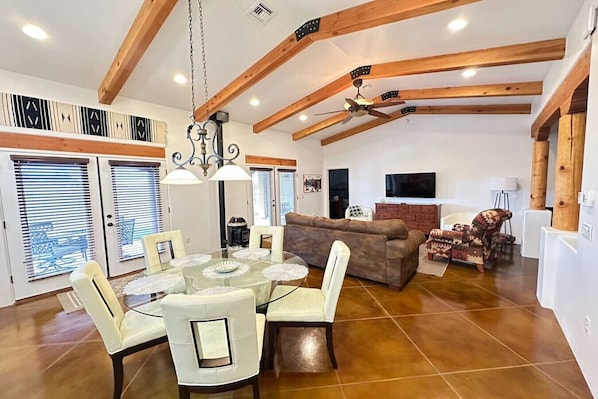 A spacious living area with a high ceiling, wooden beams, a dining table with chairs, a sofa, and a television.