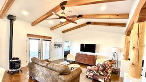 Een ruime woonkamer met een hoge plafond, houten balken, een open haard, een bank, een gestreept fauteuil, een televisie en een klein tafeltje.
