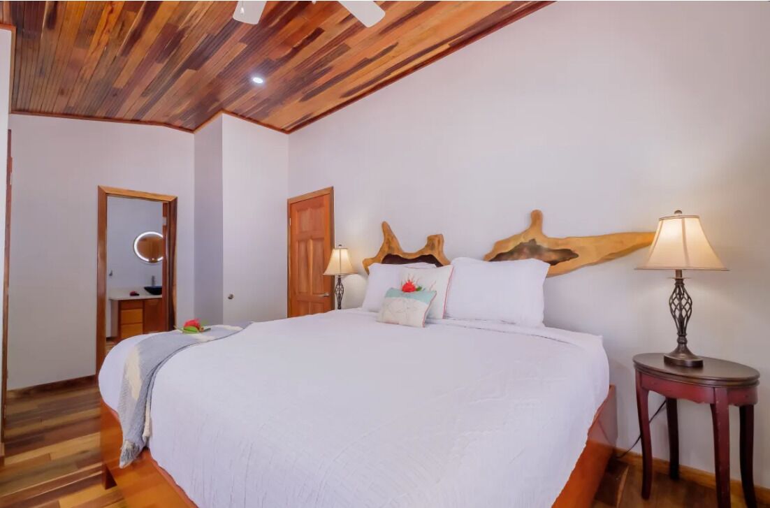Un dormitorio con una cama grande, cabecera de madera, dos mesitas de noche con lámparas, un espejo y una puerta.