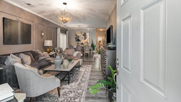 A spacious living room with a large sofa, coffee table, and a chandelier. There is a dining area with chairs and a table, and a kitchen area with cabinets and a sink.