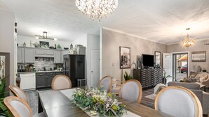 A dining area with a long table set for six, surrounded by chairs. There is a kitchen in the background with cabinets and appliances.
