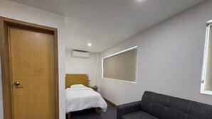 Habitació d'hotel amb una porta de fusta, un llit amb roba de llit blanca, un sofà gris i una finestra gran amb persianes.