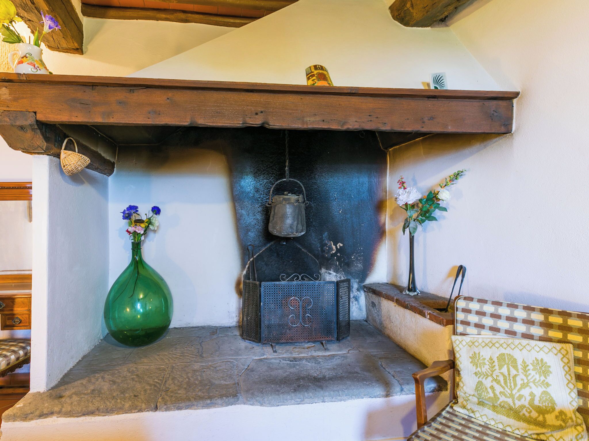 A stone fireplace with a metal pot hanging above it, a green vase with flowers on the left, and a patterned cushion on the right.