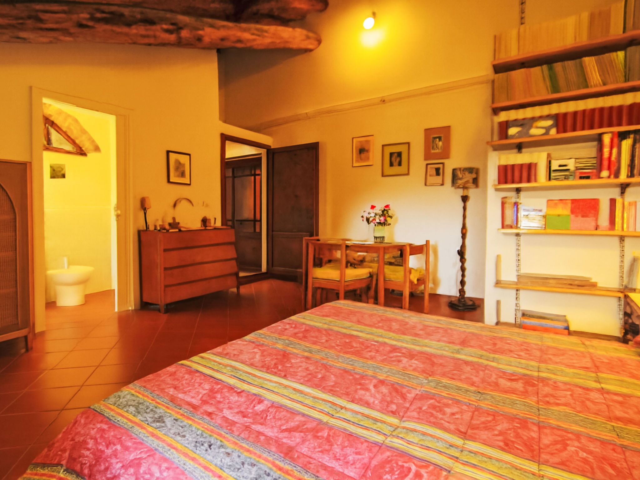 Une chambre avec un grand lit, une commode en bois, un bureau avec une chaise, une bibliothèque et une porte donnant sur une autre pièce.