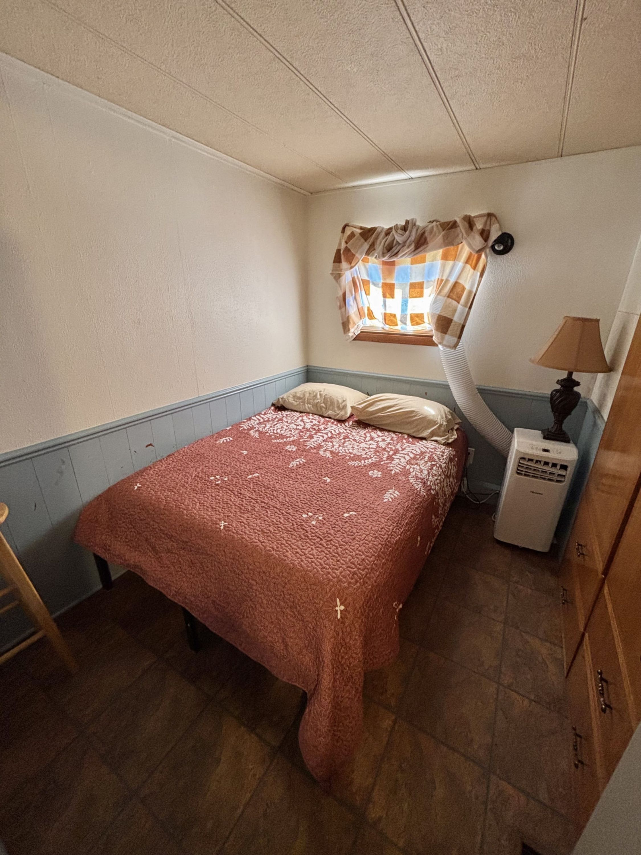 Piccola camera compatta con un letto singolo, una finestra con tenda a quadri, un comodino con lampada e un termosifone.