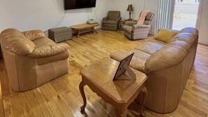 Una sala de estar con piso de madera, sofás de cuero, una mesa de centro de madera, un televisor y una ventana con persianas.