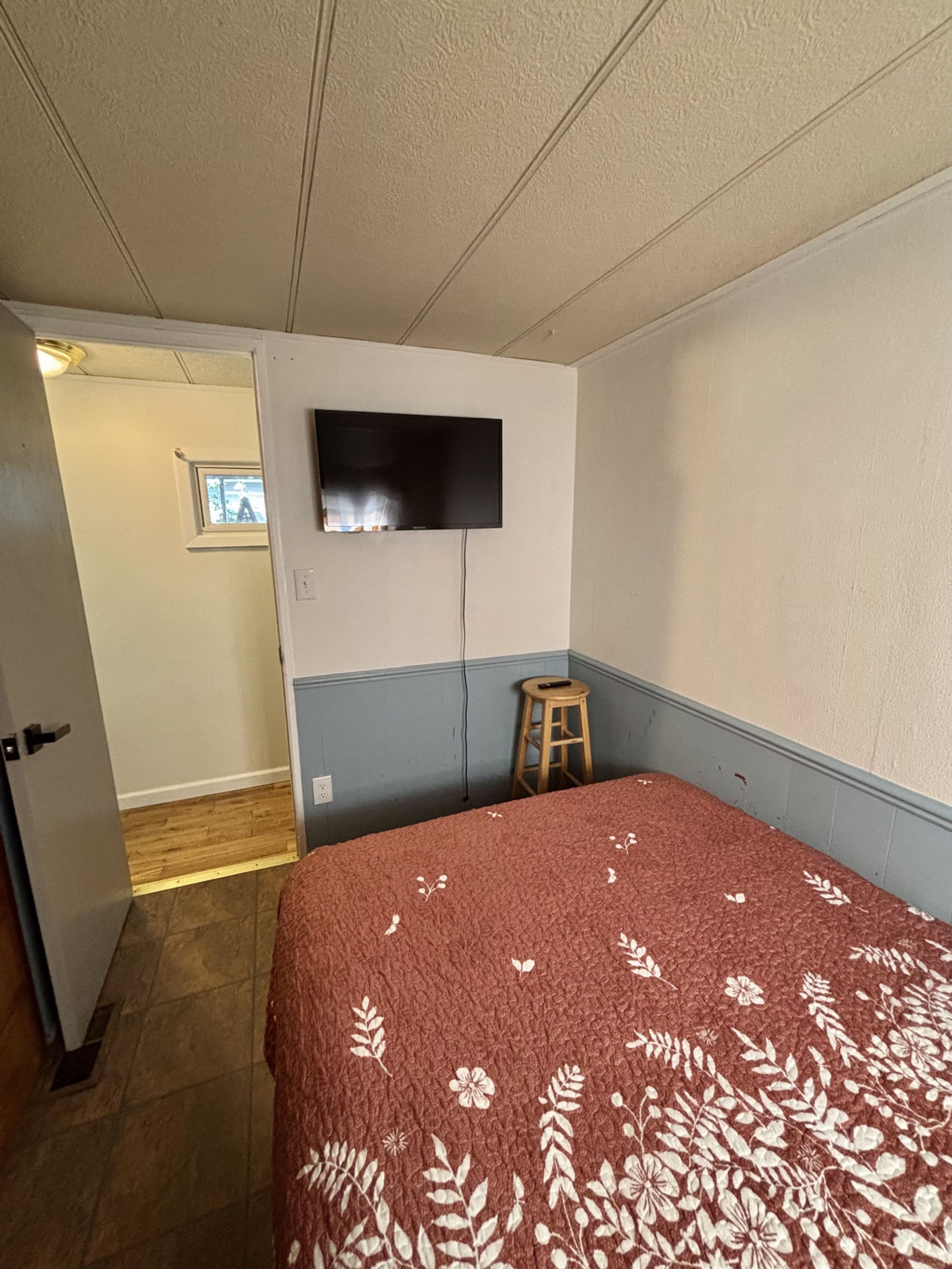 A bedroom with a bed, a television mounted on the wall, a wooden stool, and a door leading to another room.