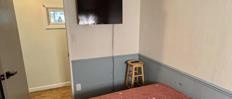 A bedroom with a bed, a television mounted on the wall, a wooden stool, and a door leading to another room.