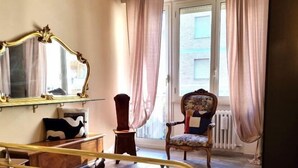 A room with a large ornate mirror, a wooden dresser, a chair with a floral pattern, and a window with curtains. - Happy Home Piramide Cestia Lussuoso Unico Esclusivo a Roma (Rome)