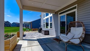 Een overdekt terras met een rieten stoel, een houten plantenbak en uitzicht op omliggende huizen.
