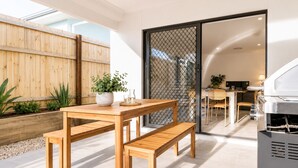 A wooden table with benches, a potted plant, and a grill in a bright, modern outdoor space. - Canopy House - Brand New Home in Nature’s Arms (Greenbank)