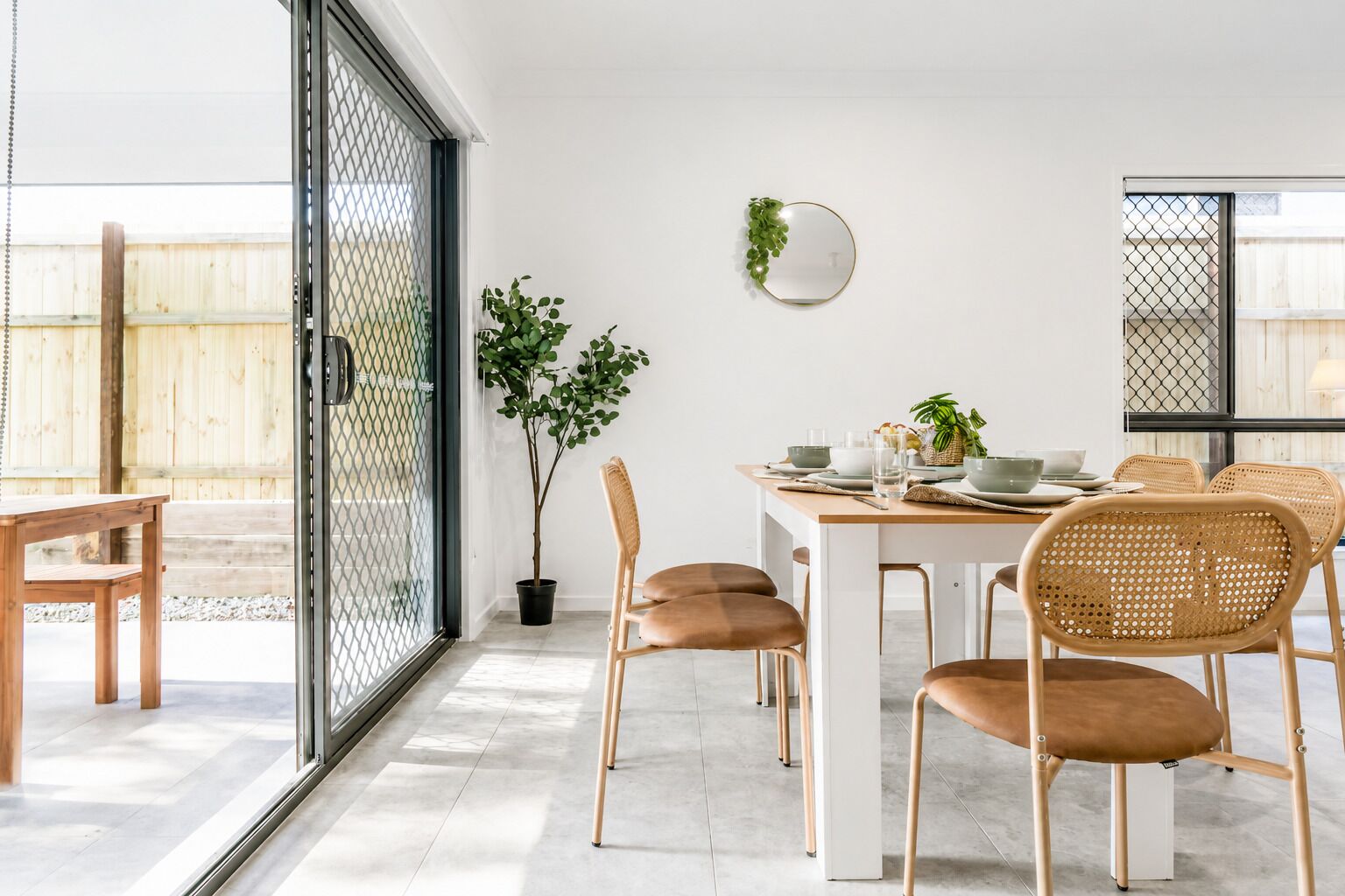 Un coin repas avec une table dressée pour quatre personnes, entourée de chaises en osier, d’une plante en pot et d’un miroir fixé au mur.