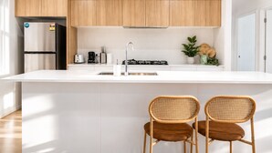 A modern kitchen with white cabinetry, a stainless steel refrigerator, a gas stove, and two wicker chairs. - Canopy House - Brand New Home in Nature’s Arms (Greenbank)