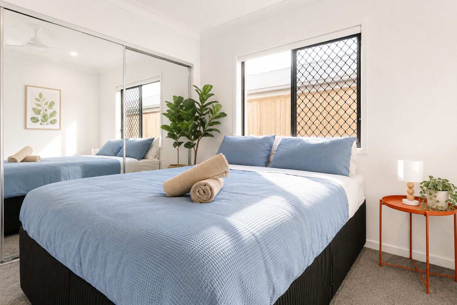 A bedroom with a large bed, a small table with a lamp, a potted plant, and a mirror.