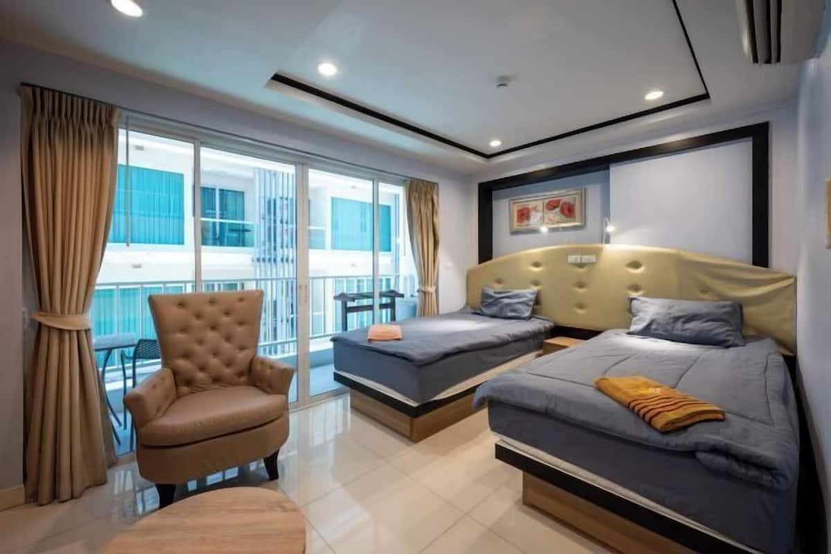 Habitación de hotel con dos camas, un sillón acolchado, una mesita y un balcón con vistas.