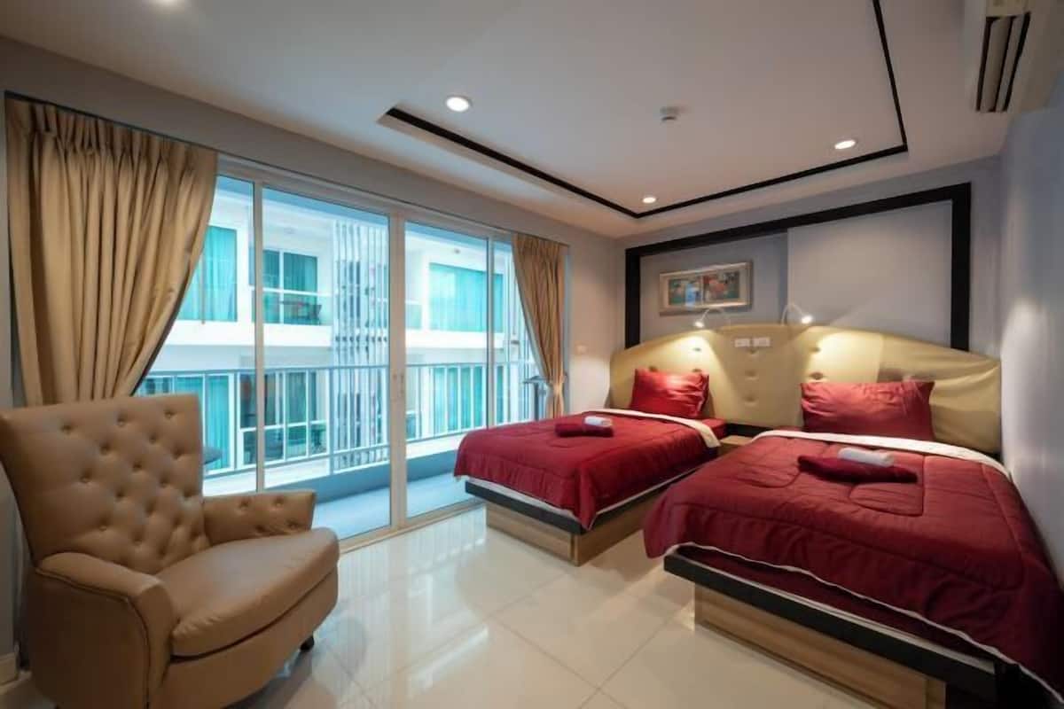 Habitación de hotel con un amplio ventanal corredero, dos camas con ropa de cama roja, un sillón color beige e iluminación empotrada en el techo.