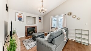 Ein modernes Wohnzimmer mit einem grauen Sofa, einem Kamin, einer Chandelüster und einem Fenster mit Vorhängen.