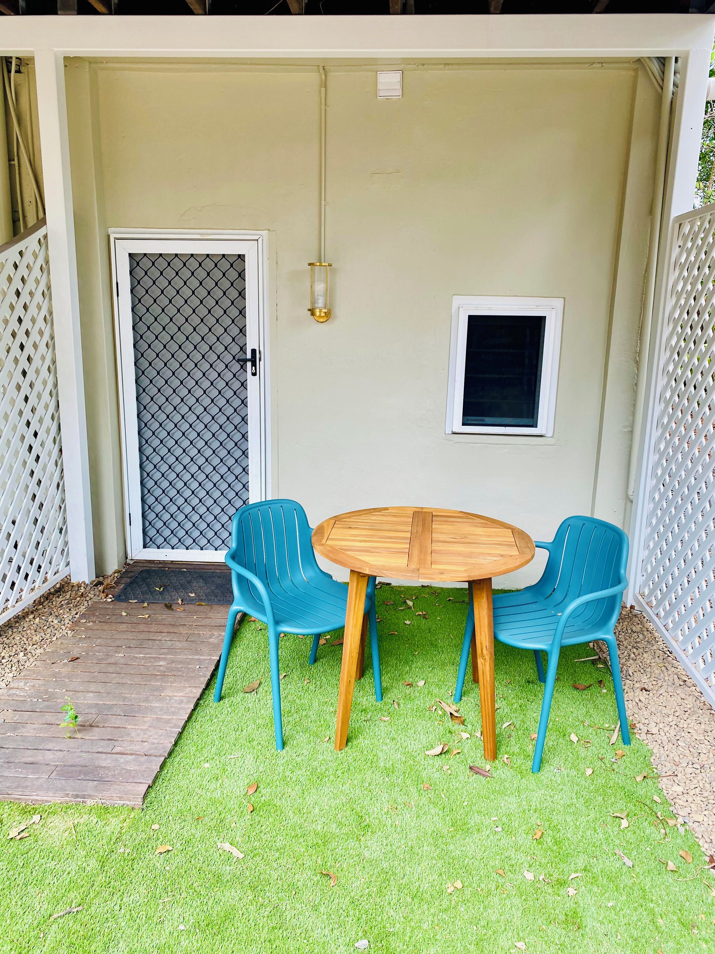A covered patio with a wooden table and two blue chairs, a door with a mesh screen, and a small window.