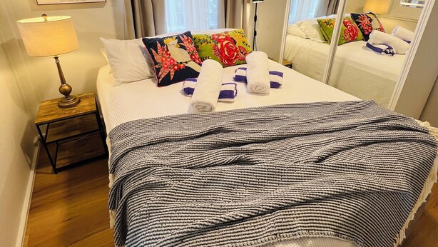 A hotel room with a bed, a nightstand, a lamp, a mirror, and a window with curtains. - City Unit 4 Walk to Esplanade (Cairns North)