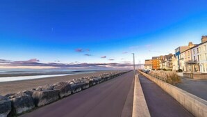A coastal road with a stone barrier, a clear sky, and buildings on the right. - Sunbeam House Apartment 3 (Morecambe)