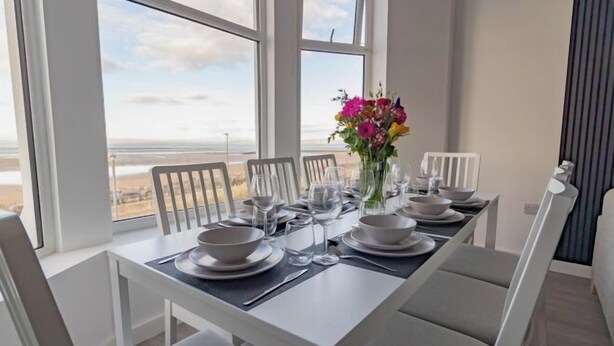 A dining table set for six with white chairs, a vase of flowers, and a view of the beach. - Sunbeam House Apartment 3 (Morecambe)