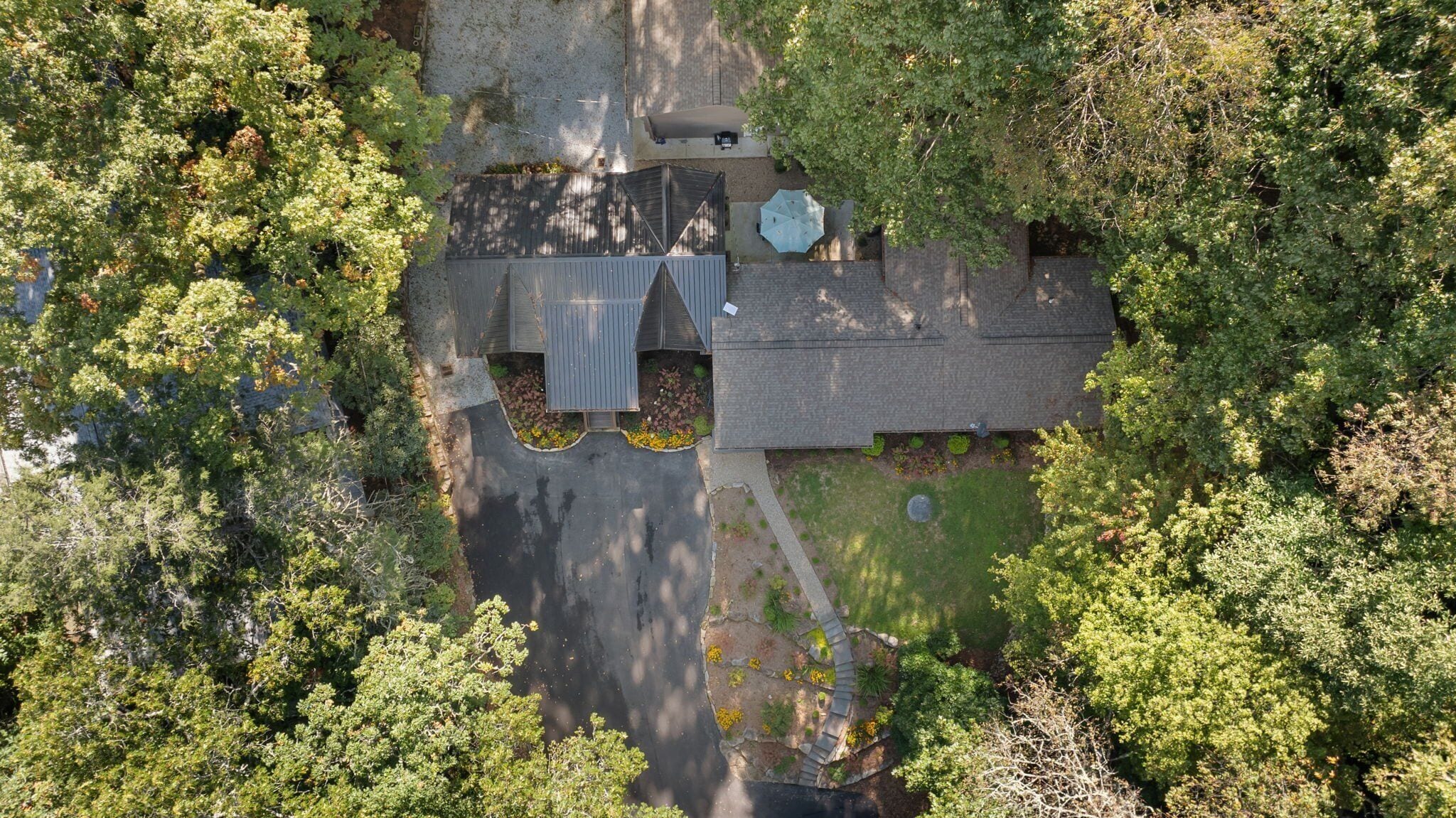 Une vue aérienne d’une maison entourée d’arbres denses, avec une allée menant à un garage et une terrasse couverte.