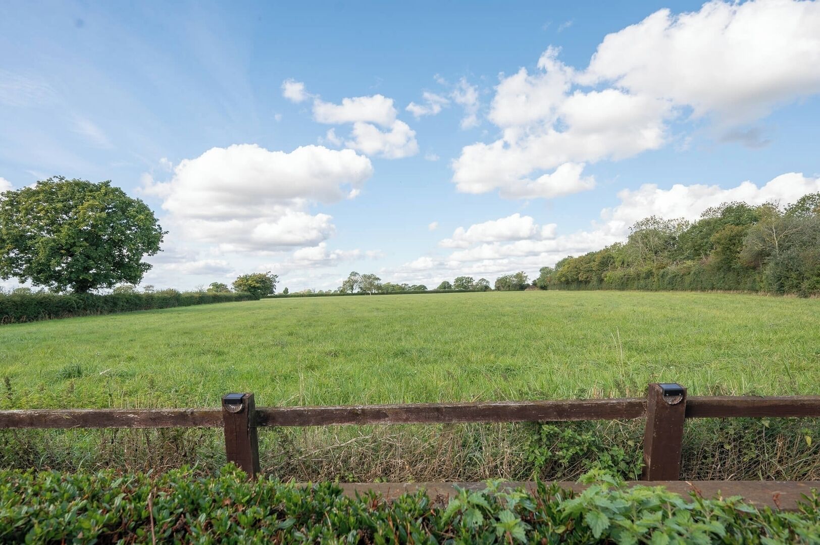 A grassy field with a wooden fence in the foreground and trees in the background under a partly cloudy sky.