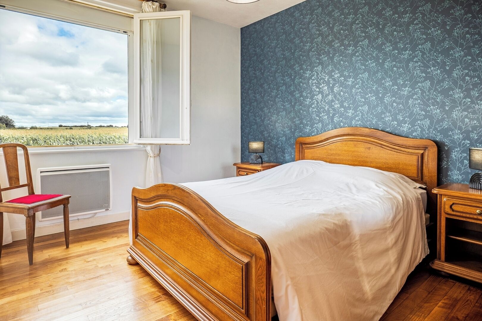 A bedroom with a wooden bed, a chair, a nightstand, and a view of a field through a window.