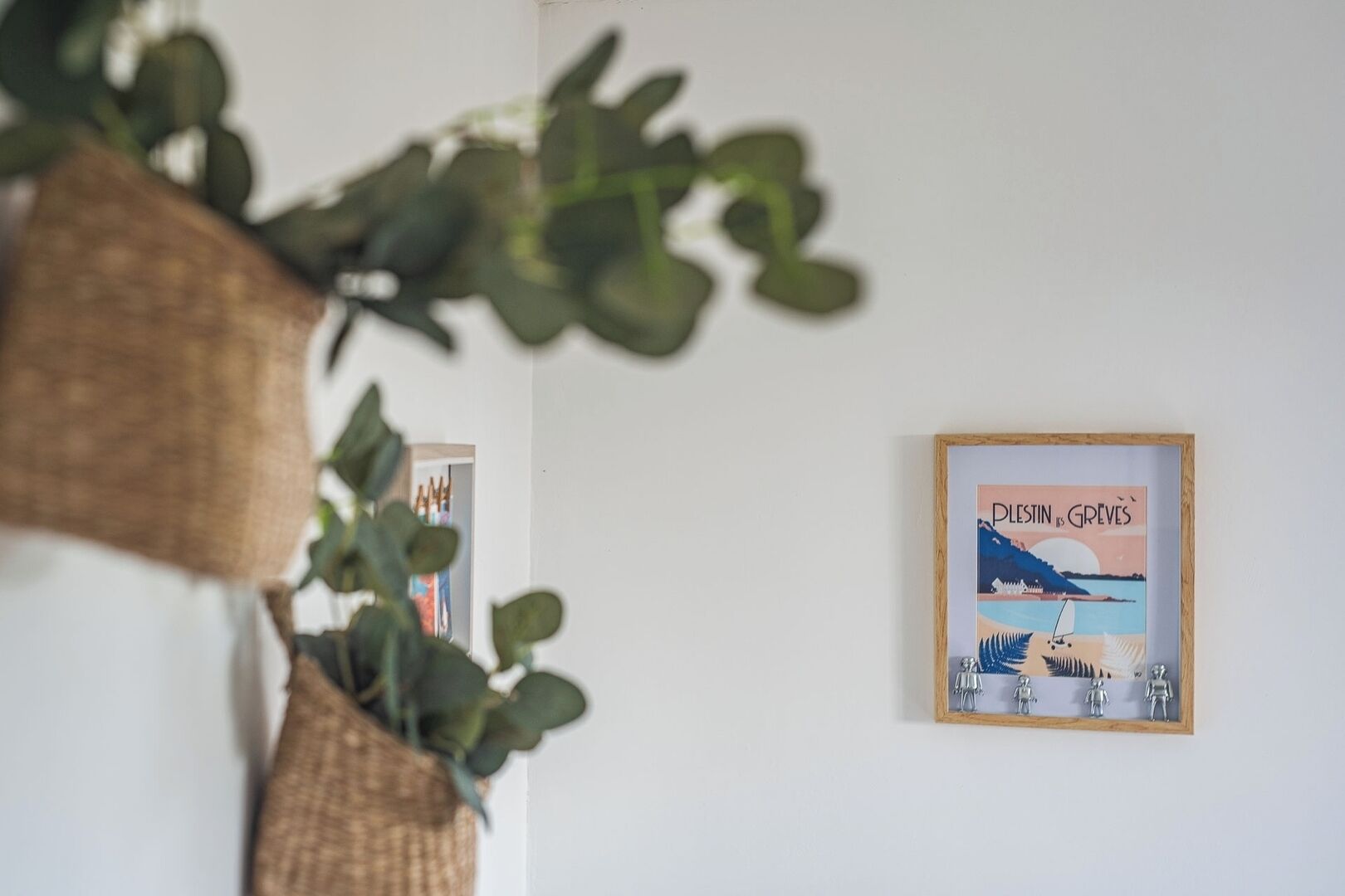 Una obra de arte en marco con una escena costera y texto, una planta colgante y un vistazo a una habitación con una puerta y una pared.