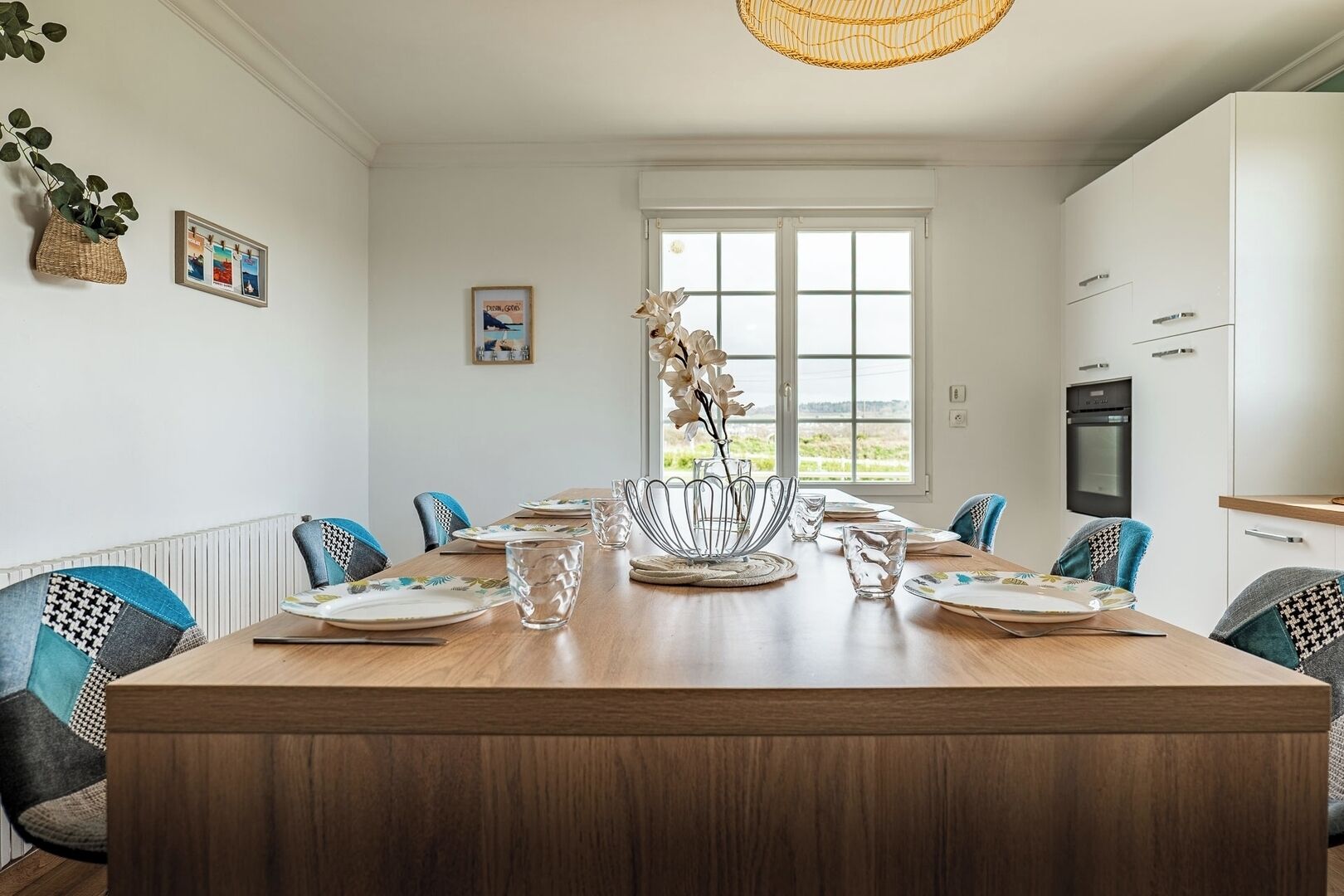 A dining area with a large wooden table, chairs, and a vase of flowers. There are framed pictures on the wall and a window with a view of the outdoors.