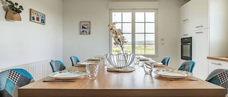 A dining area with a large wooden table, chairs, and a vase of flowers. There are framed pictures on the wall and a window with a view of the outdoors.