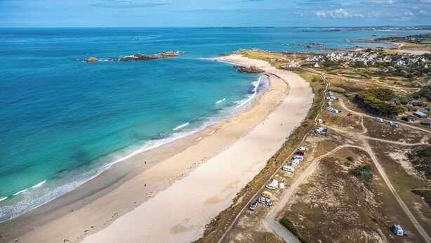 Una vista aerea di una zona costiera con spiaggia sabbiosa, un piccolo insediamento con case e veicoli parcheggiati, e un cielo azzurro limpido.