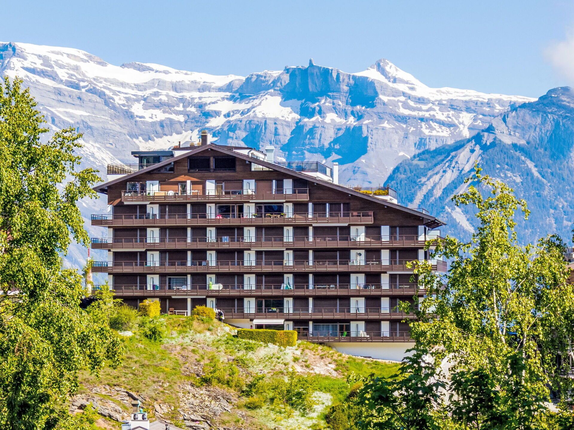 A multi-story hotel with balconies, set against a backdrop of snow-capped mountains.
