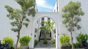 A courtyard with a white archway, surrounded by potted plants and trees, leading to a building with a modern design. - Inori Villas (Bali)