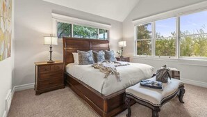 A bedroom with a large bed, two bedside tables, a chair, and a window with a view of trees.
